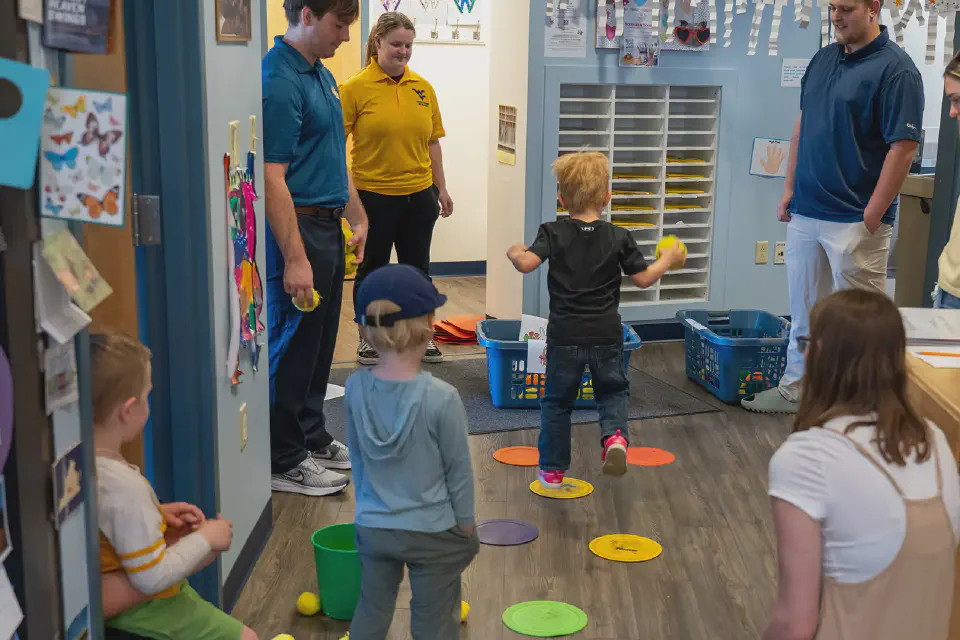 A preschooler jumps across colorful floor markers during an indoor activity, thoughtfully observed by WVU students and teachers.