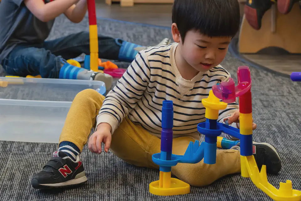 A young boy sits on a classroom rug, focused on building a colorful marble run while another student plays in the background.