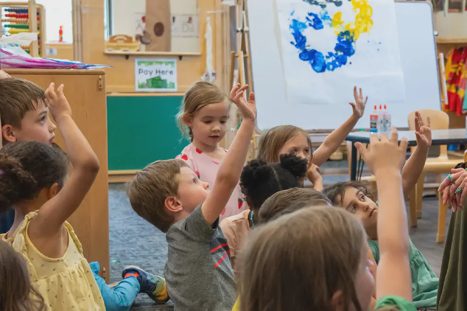 Diverse preschoolers sit on the classroom floor, eagerly raising their hands to participate in a lesson with a smiling teacher.